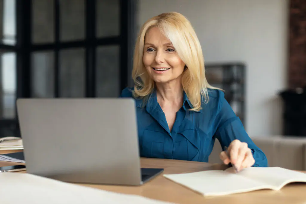 Remote work. Smiling mature woman entrepreneur using laptop at home, looking at computer screen smiling and taking notes