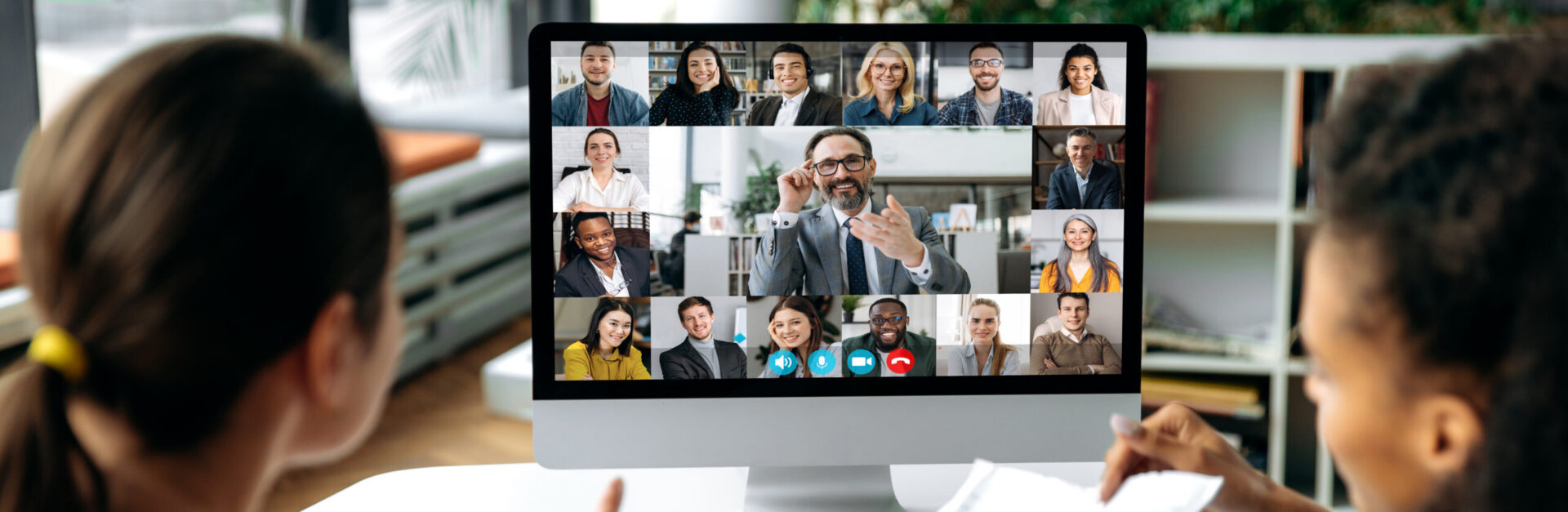 Virtual meeting online, video call. View over shoulders of two women to a computer screen with business leader and successful team, chatting by a video conference, discuss working issues, strategy