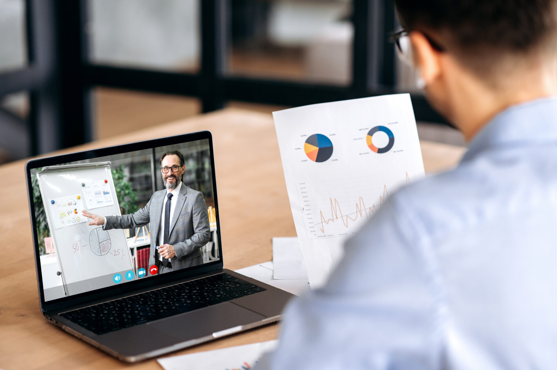 Over shoulder view of a caucasian male manager holding financial graphs, having online video conference with mature successful ceo company using laptop, sitting in office, concentrated