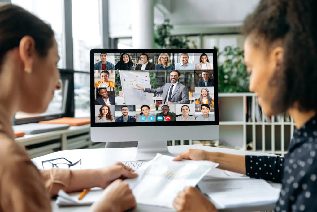 Online business meeting, brainstorm. View over the shoulders of two women to a computer screen with different successful multiracial people gathered in a video conference to discuss working issues