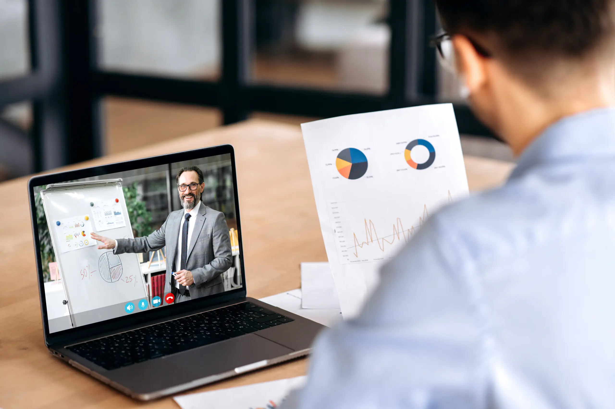 Over shoulder view of a caucasian male manager holding financial graphs, having online video conference with mature successful ceo company using laptop, sitting in office, concentrated
